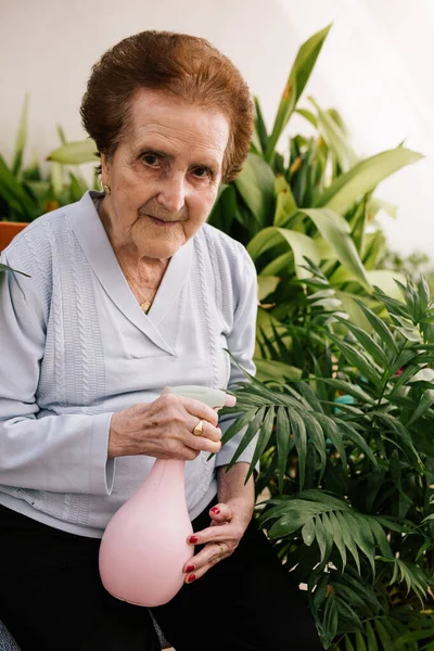 Terasındaki çiçekleri sulayan yaşlı bir kadının portresi.