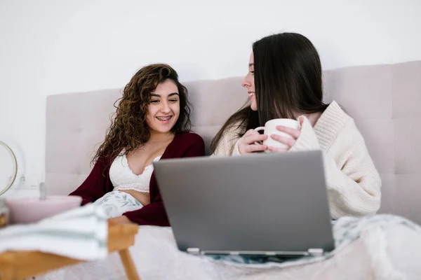 Yatakta iki beyaz kadın dizüstü bilgisayara bakarken biri elinde bir bardak tutuyor.