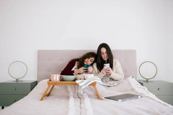 Lezbiyen çift kahvaltı yaparken cep telefonuna bakıyor. Yatağın üzerinde kahvaltı masası ve dizüstü bilgisayar var.