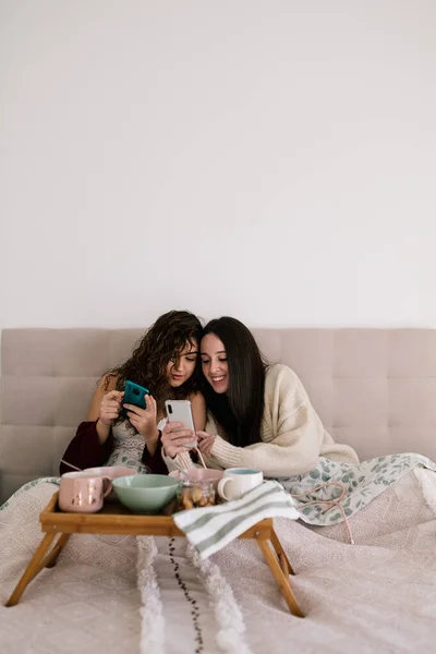 İki tane siyah saçlı beyaz kadın cep telefonuna bakarken yatakta uzanıyorlar. Küçük bir masada kahvaltı hazırlıyorlar.