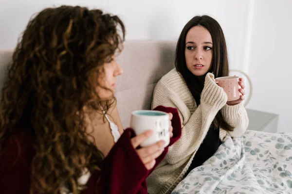 İki beyaz kadın bir yatakta birbirlerine bakarken kupa tutuyorlar.