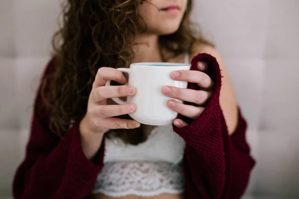 Yatakta esmer saçlı beyaz bir kadının portresi. İki eliyle bir kupa tutuyor.