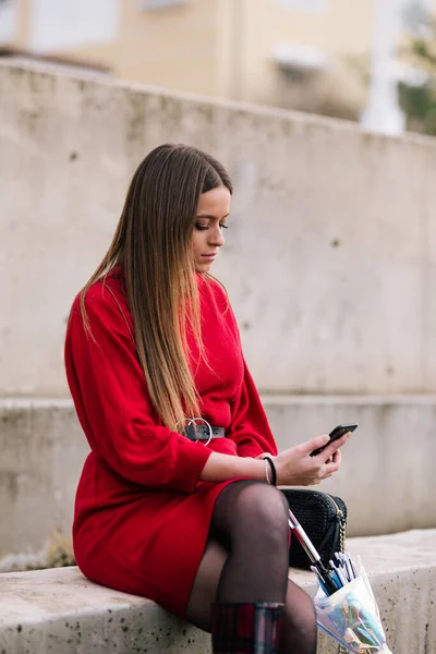 Kumral saçlı, kırmızı elbiseli, açık havada oturan ve cep telefonuna bakan genç bir kadın.