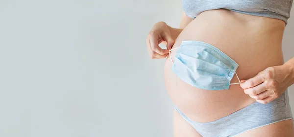 Gri arka planda tıbbi koruyucu maskesi olan hamile bir karın. Koronavirüs salgınında gebelik kavramı, karmaşık hamilelik, annelik, tüp bebek, covid-19 salgını, toksikoz