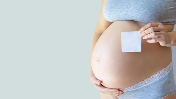 Pregnancy promo. Mockup white sheet in hand of pregnant woman on gray background. Concept pregnancy, motherhood, love, In vitro fertilization, abortion, toxicosis, IVF, family and single mother