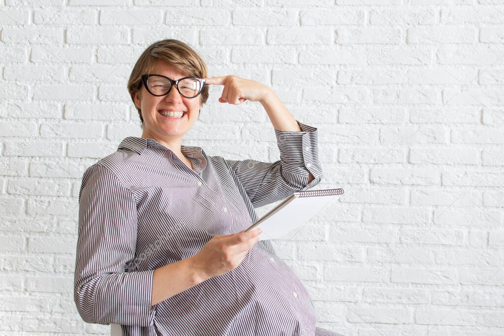 Young pregnant woman in glasses with a notepad in her hands and a finger to her temple. Pregnant woman working concept, planning shopping, childbirth, sitting at her working place in office. Copyspace