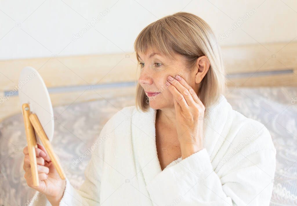 Senior woman in the bedroom applying anti-aging lotion. Looks in a cosmetic mirror. Concept Anti age, healthcare and cosmetology, old age, pensioner and mature