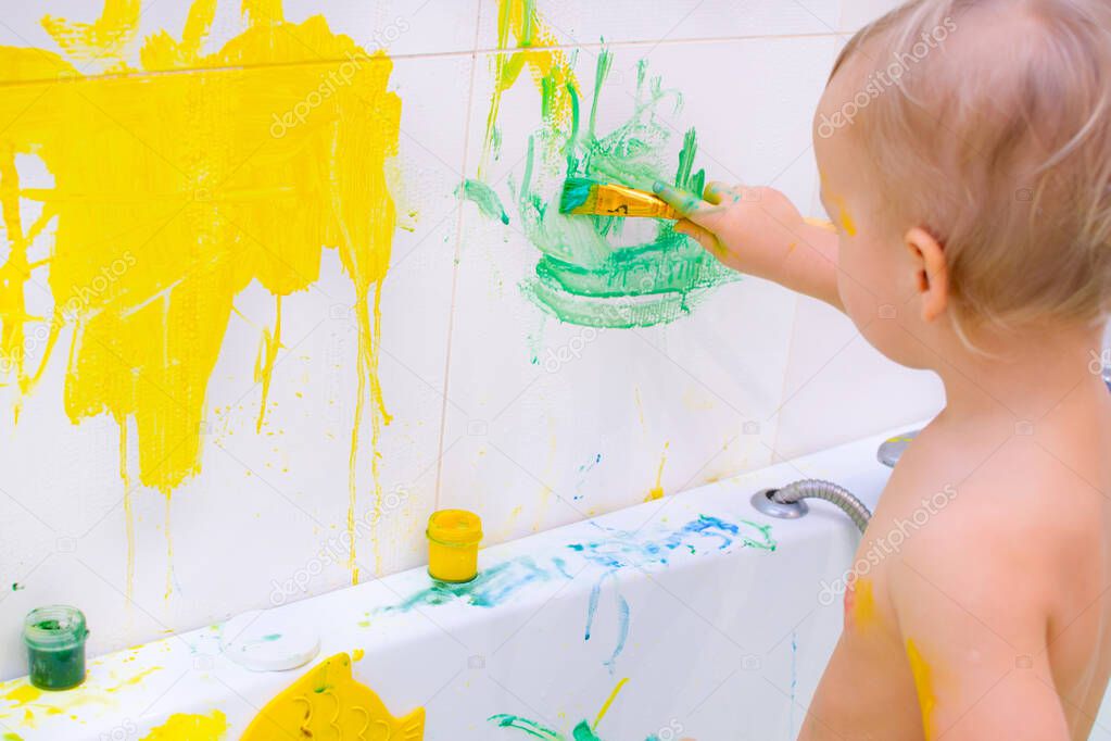 Kid draw with paints in the bath, playing at home. Todler with educational toys for creativity. Boy play at home. Bright paint gouache stained face and hands of a child.