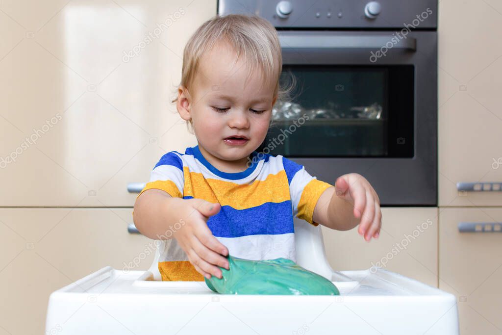 Child play with slime at home in the kitchen. Little toddler boy squeeze and stretching green slime sits at a kids table. Child having fun and being creative plays