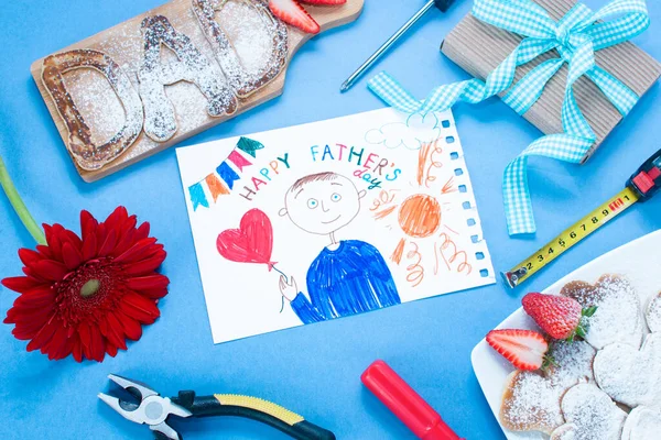 Father day Breakfast pancakes DAD letters, gift, building tools on a blue background. Childs drawing of her dad. Concept Happy Fathers Day flat lay