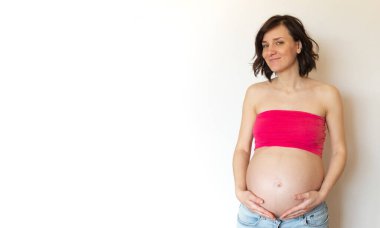 Hafif arka planda hamile gerl afişi. Kot pantolonlu, gülümseyen hamile bir kadın karnına sarılıyor. Anneliğin neşesi ve mutluluğu. Kopyalama alanı