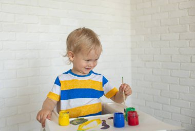 Resimli çocuk çizimi, evde oynamak, çocuk masasında oturmak. Yaratıcılık için eğitici oyuncakları olan bir çocuk. Çocuk evde oynuyor. Yüzü ve elleri parlak boya lekeli bir çocuk. Çocuk eğleniyor.
