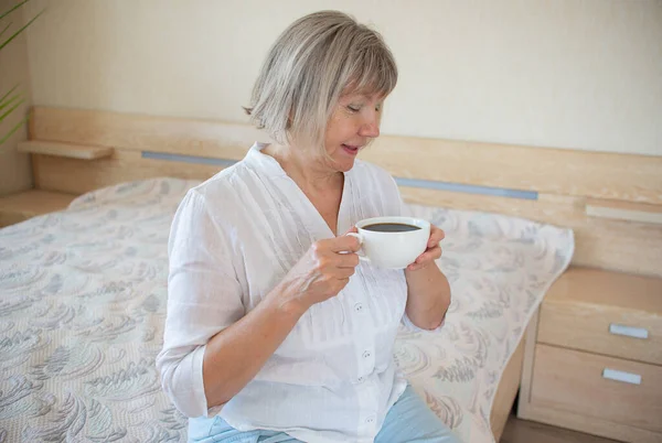 Старша жінка тримає чашку кави, розслабляючись, посміхаючись у своєму будинку в спальні. Доброго ранку прокидається концепція — стокове фото