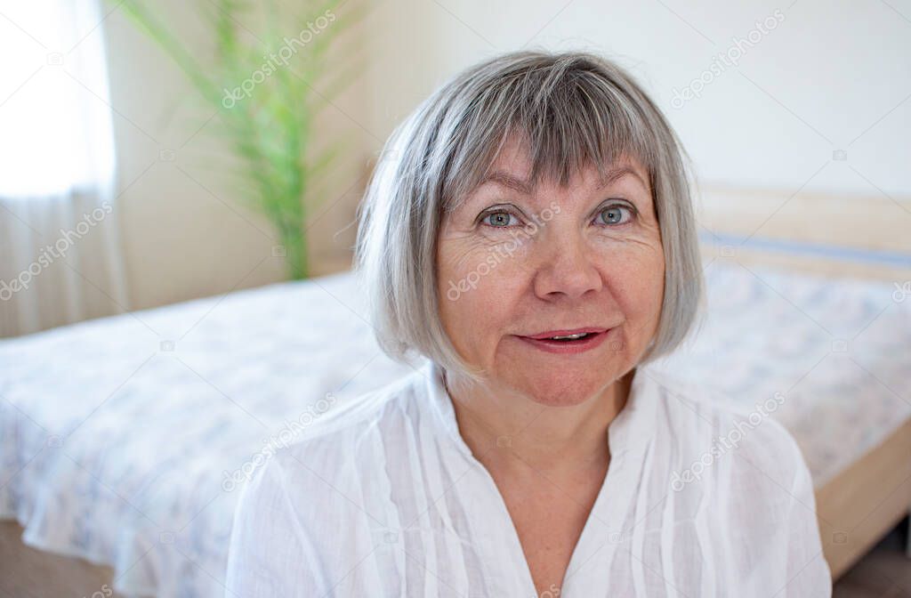 Happy senior woman with gray hair relaxing smiling is looking at the camera in her home in the bedroom.