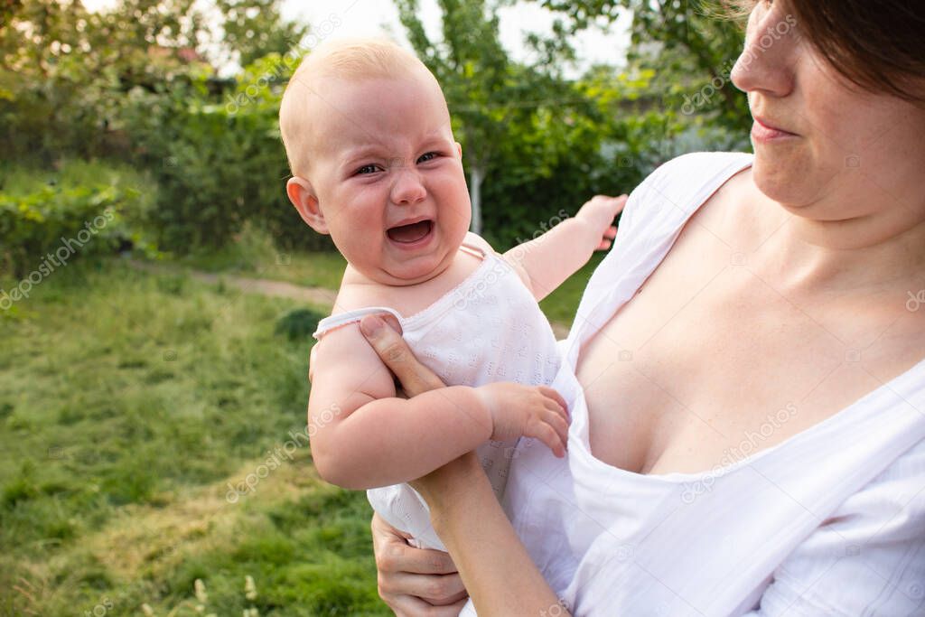 Crying little blonde kid crying in moms arms outdoors in the garden outdoors