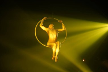 Esnek genç bir kadın hava çemberinde, sirk gösterisinde, sarı ışıkta performans sergiliyor. Esnek kadın jimnastikçi, siyah arka planda oturuyor.