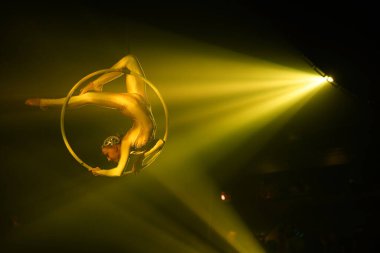 Esnek genç bir kadın, hava çemberinde performans sergiliyor, hava çemberinde esnek, sirk gösterisi, sarı ışık. Esnek kadın jimnastikçi potada baş aşağı duruyor. Altın kostüm, sağda boş alan