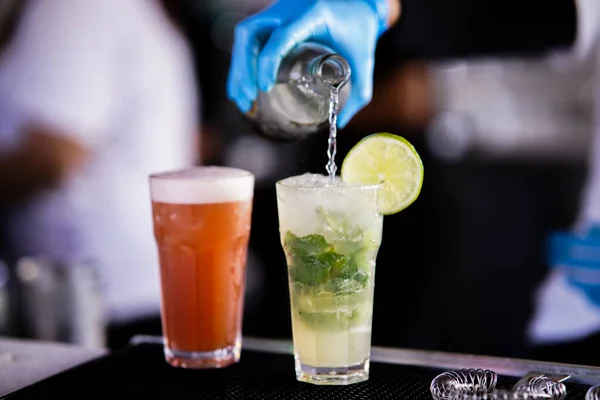 A glass of grapefruit juice, the bartender pours soda into a mojito, hands with gloves