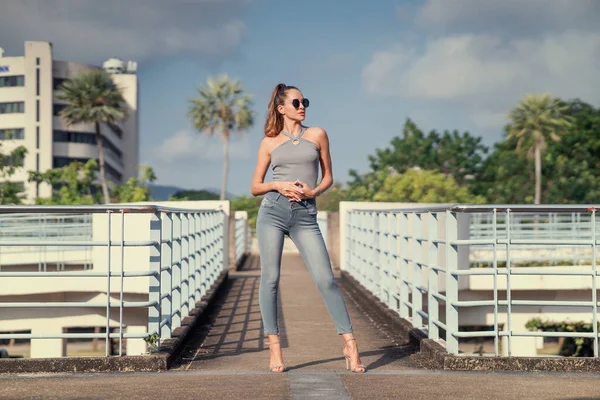 Tayland 'da kadın model, gri kot pantolon, gri bluz, güneş gözlüğü, yaz zamanı, arka plan palmiyeleri ve şehirle moda filmi. Yüksek kalite fotoğraf