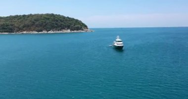 Beyaz büyük yat yeşil tepeler ve küçük gemilerle çevrili pitoresk bir körfezde duruyor. Kamera soldan sağa hareket ediyor.