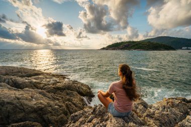 Genç bir kadın deniz kenarında meditasyon yapıyor. Lotus pozisyonunda oturuyor. Denize bakan kayaların üzerinde.