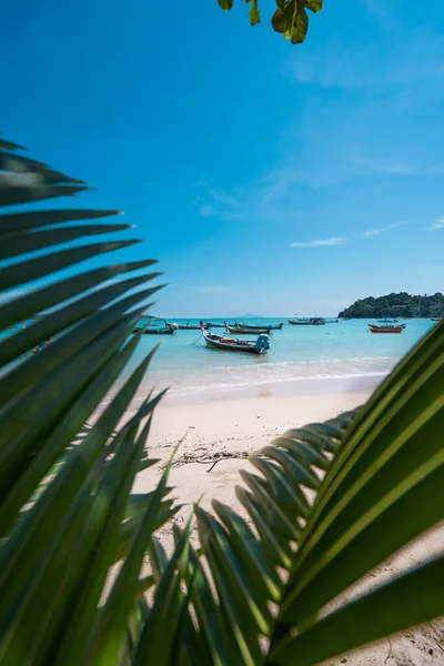 Thailändisches Fischerboot an der türkisfarbenen Küste, eingerahmt von Palmenblättern. Sommerurlaub und Naturerlebnis-Konzept. Vertikales Foto — Stockfoto