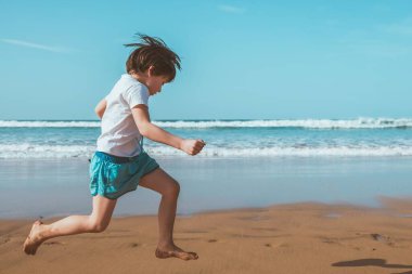 5 yaşında bir çocuk elinde mavi bir kovayla plajda koşup zıplıyor.