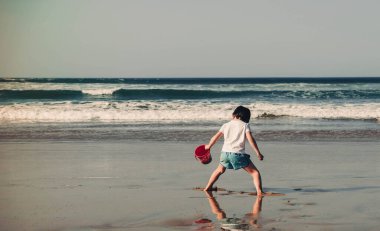 Elinde mavi bir kovayla plajda oynayan 5 yaşında bir çocuk.