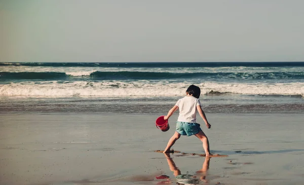 Elinde mavi bir kovayla plajda oynayan 5 yaşında bir çocuk.