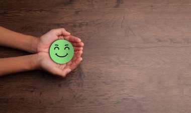 Hands gently holding a green happy face symbol on a wooden table background, representing positivity, emotional support, and mental health and wellness with space for text on the right.
