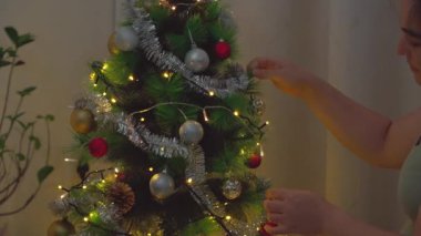 Genç bir kadın Noel ağacını güzel bir evde süslüyor. Yumuşak ışıklar, mevsimlik süsler ve neşeli bir atmosferle dolu sıcak ve neşeli bir bayram anı. C temaları için mükemmel
