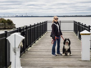 Nehre bakan ahşap bir iskelede duran siyah ve beyaz bernedoodle köpeğiyle su boyunca endüstriyel rafinerisi olan bir kadın..