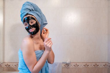 Genç bir kadın yüzünde siyah bir maske ve kafasında bir havluyla banyoda gülümsüyor. Sabah rutini boyunca cilt bakımı kavramı.