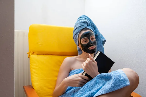 Young woman sitting on the couch with a book on her hands, a black facial mask and a towel rolled up on her head. Concept of skin care during the morning routine.