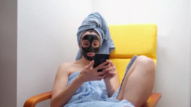 黒い顔のマスクとタオルを頭に巻いて携帯電話で自画像を撮る若い女性 朝の習慣の間のスキンケアの概念 — ストック動画
