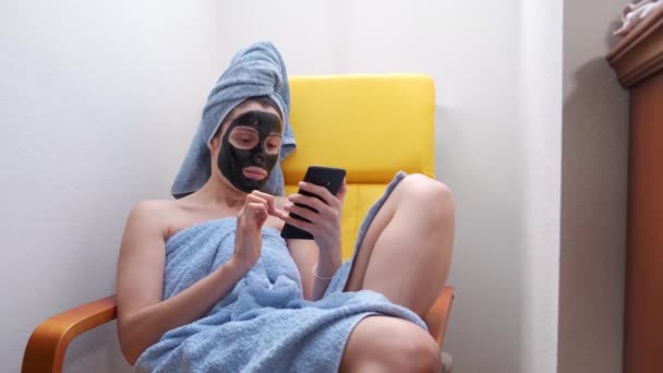 黒い顔のマスクとタオルでソファの上に携帯電話を使用して若い女性が彼女の頭の上にロールアップ 朝の習慣の間のスキンケアの概念 — ストック動画