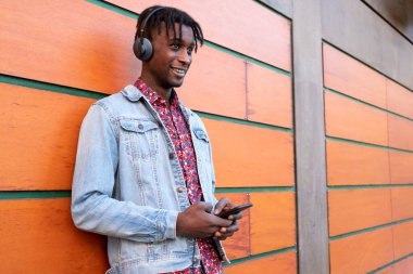 Genç Afrikalı adam ahşap arka planda kulaklıkları ve akıllı telefonuyla gülümsüyor..