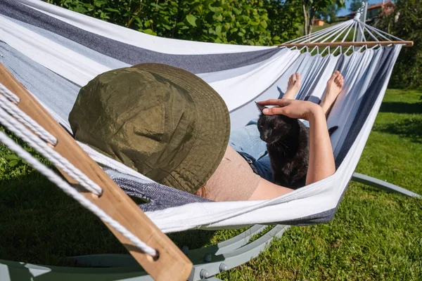 Relaxed woman in hammock with a black cat pet enjoying good weather in the garden. She is wearing a bucket hat.