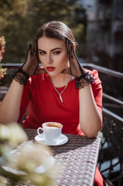 Kafede oturmuş, bir fincan kahve içen genç ve şık bir kadın. Kırmızı elbise ve siyah eldiven giyen esmer kadın. Şehir sokağı, Avrupa tatili. Zarif kadın.