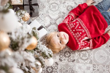 6 yaşında kırmızı kazaklı bir çocuk Noel ağacının altında topları ve çelengi ile yatıyor. Tatlı sarışın çocuk.