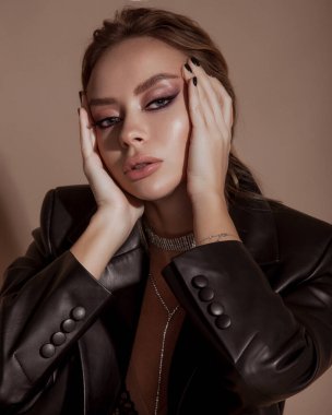 Yakın plan güzellik portresi. Parlayan sağlıklı cildi olan genç bir manken. Parlak makyajlı güzel sarışın kadın. Deri ceket giyen ve bej arka planda poz veren bir manken.