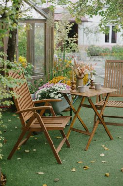 Ahşap bir katlama masası ve iki sandalye arka planda çiçeklerle dolu yeşil bir çimenlikte oturuyorlar. Bu da sonbahar bahçesinde inzivaya çekildiğini gösteriyor..