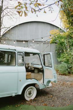 Klasik açık mavi minibüs, yaprakları dökülen ahşap bir ambarın önüne park edilmiş..