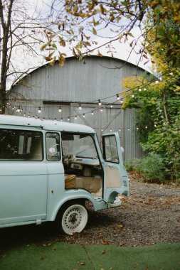 Klasik açık mavi minibüs, yaprakları dökülen ahşap bir ambarın önüne park edilmiş..
