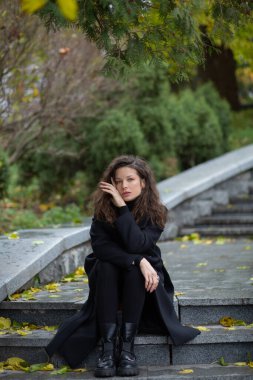 Taş merdivenlerde oturan düşünceli genç bayan. Sonbahar parkında. Siyah ceketli kadın.