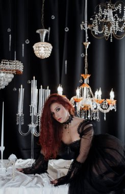 Uzun kızıl saçlı, siyah elbiseli, zarif bir odada kristal avizelerle poz veren güzel bir kadın.