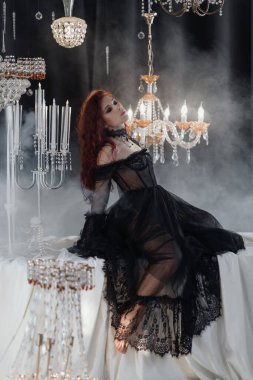 Uzun kızıl saçlı, siyah elbiseli, zarif bir odada kristal avizelerle poz veren güzel bir kadın.