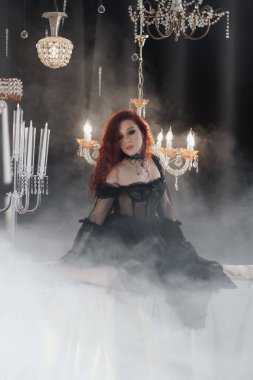 Uzun kızıl saçlı, siyah elbiseli, zarif bir odada kristal avizelerle poz veren güzel bir kadın.