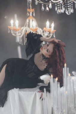 Uzun kızıl saçlı, siyah elbiseli, zarif bir odada kristal avizelerle poz veren güzel bir kadın.