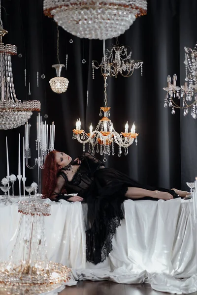 Uzun kızıl saçlı, siyah elbiseli, zarif bir odada kristal avizelerle poz veren güzel bir kadın.